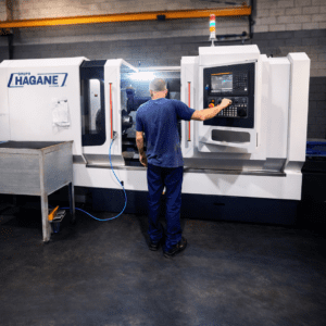 Um técnico industrial opera um torno mecânico de grande porte durante o serviço de recuperação de rolos de conformação. Faíscas saem do ponto de contato da ferramenta com o metal. O técnico veste uma camisa azul com o logotipo branco do Grupo Hagane nas costas.